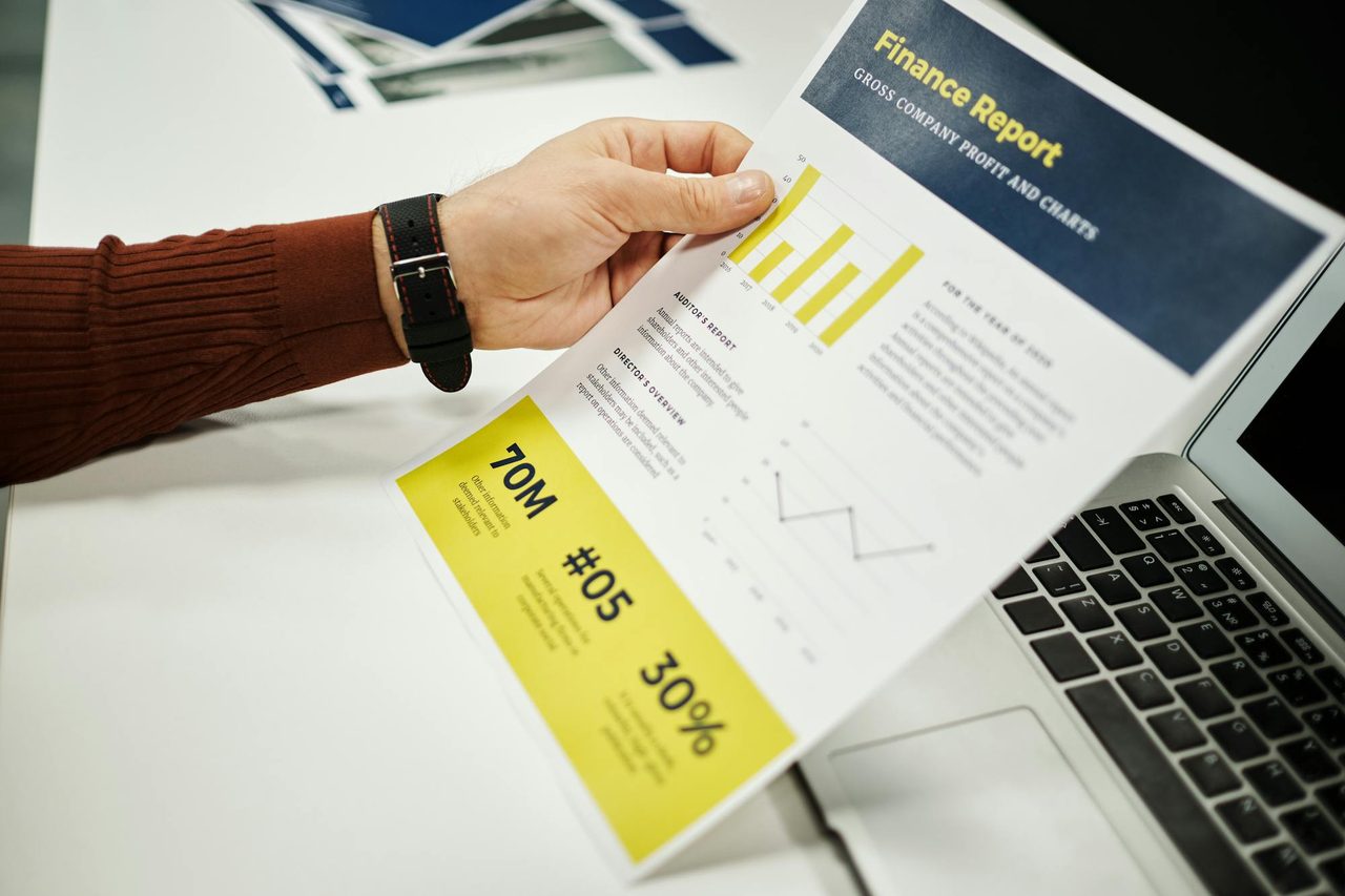 A hand holding a printed finance report with charts beside a laptop